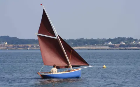 Un voilier bleu avec une voile marron navigue, tel une gazelle des îles, sur une mer calme. Au loin, une côte boisée bordée de maisons.
