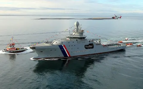 Un imposant bateau garde côte, le Landhelgisgæslan, navigue en mer, escorté par un remorqueur et suivi par des canots. Un hélicoptère survole la scène.