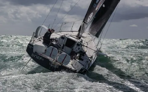 Xavier Macaire navigue sur un voilier dans une mer agitée, le bateau est incliné, les vagues déferlent.