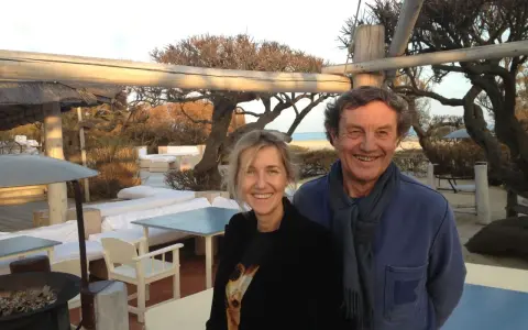 Patrice de Colmont et une femme souriants sur une terrasse de plage, avec des arbres dénudés et des sièges blancs en arrière-plan.