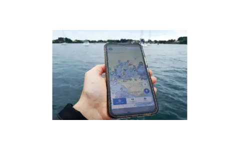 Main d'une personne tenant un smartphone affichant une carte marine avec des points d'intérêt. Le nav et co. sont visibles sur l'eau.
