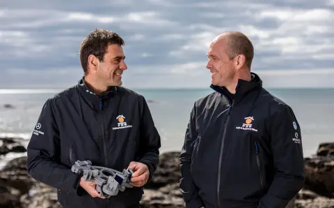 Damien Guillou et son collègue souriants sur la plage, l'un tenant un sextant, prêts pour une nouvelle aventure maritime.