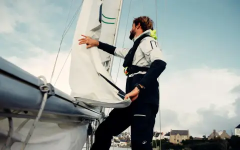 Un homme ajuste un type de voile sur un voilier, avec des maisons en arrière-plan.