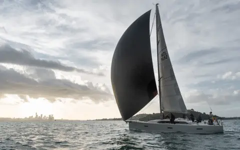 Un voilier navigue avec un grand gennaker noir gonflé par le vent, contrastant avec le spinnaker gris. La ville d'Auckland se dessine à l'horizon.