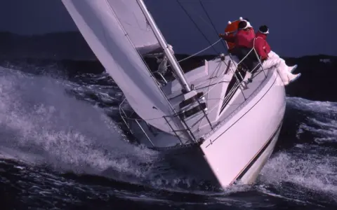 Voilier blanc fendant les vagues, deux personnes en ciré rouge. Une scène de navigation intense, digne des premiers 285 avis.
