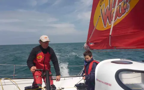 Franck-Yves Escoffier et un jeune garçon naviguent sur un voilier par temps ensoleillé.