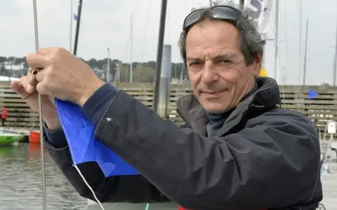 Marc Guillemot, skipper expérimenté, ajuste une voile bleue sur un ponton, prêt pour une nouvelle aventure en mer.