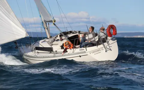 Un bateau symphonie navigue sur des eaux bleues, trois personnes à bord profitent du soleil.