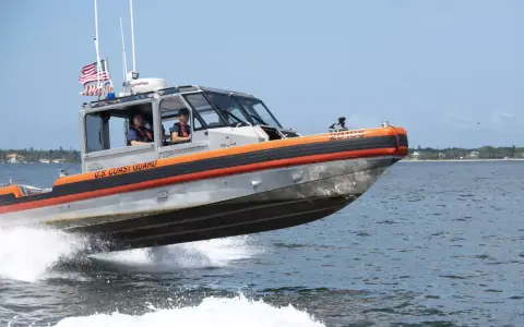Un bateau du garde côte américain file sur l'eau, créant une gerbe d'écume. Deux membres d'équipage sont visibles à l'intérieur.
