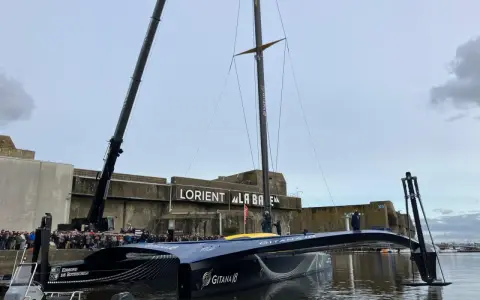 Le trimaran Gitana 18, prêt pour l'aventure, est soulevé par une grue à Lorient. Une foule nombreuse est rassemblée pour admirer ce géant des mers.