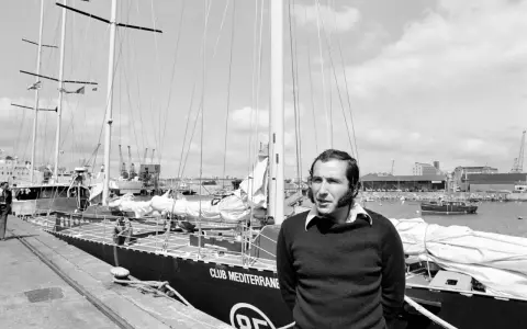 Un homme pose devant le voilier "Club Mediterranee". Ce navigateur célèbre semble prêt pour une nouvelle aventure maritime.