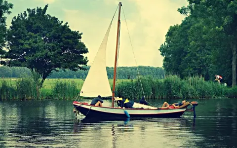 Un petit type de bateau à voile navigue sur un canal bordé d'arbres. Une personne se repose sur le pont, une autre est assise.