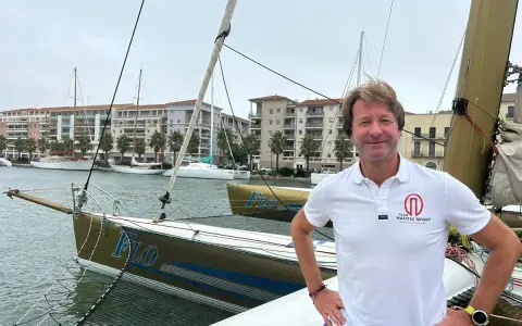 Un homme souriant sur un trimaran, peut-être le "Florence Arthaud", avec des bateaux et des immeubles en arrière-plan.