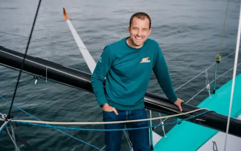 Sam Goodchild, souriant, sur un voilier, portant un sweat "For the Planet".