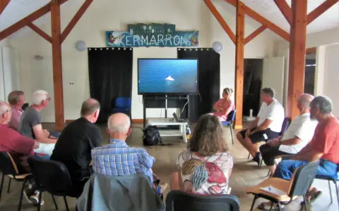 Gwen Chapalain, une femme assise, regarde un écran diffusant une image de bateau. Un groupe écoute attentivement.