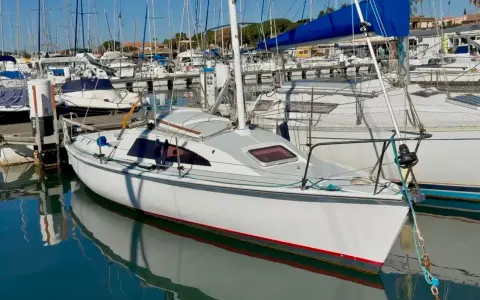 Un voilier blanc avec une voile bleue dans un port, reflété dans l'eau calme.
