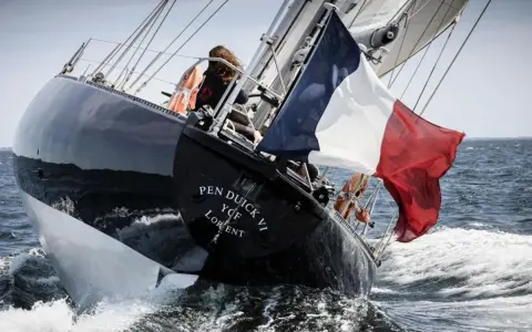 Le voilier Pen Duick VI fend les vagues, pavillon français au vent. Une silhouette se tient à la barre.