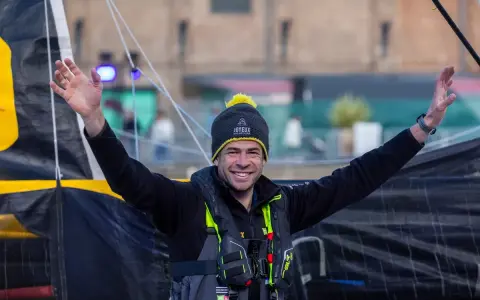 Nicolas d'Estais, marin souriant, bras levés, porte un bonnet et un gilet de sauvetage, prêt pour l'aventure en mer.