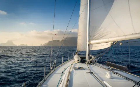 Le voilier navigue sur une mer bleue, le grand voile blanc gonflé par le vent. Au loin, des îles brumeuses se dessinent sous un ciel clair.