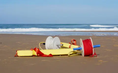 Deux bobines de fil, une blanche et une rouge, sont attachées à un tube de sauvetage jaune sur une plage.