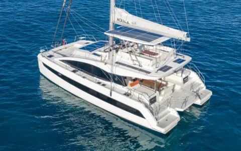 Un catamaran blanc, une véritable bicoque bateau, navigue sur une mer bleue. Ses voiles sont partiellement déployées, et des panneaux solaires brillent sur son toit.