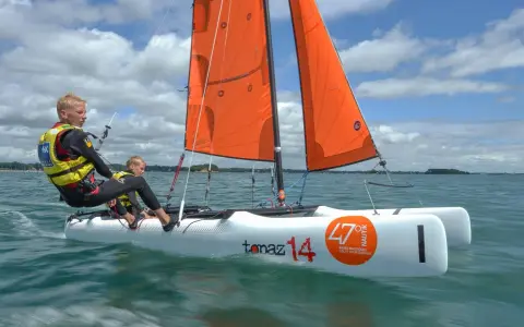 Deux jeunes garçons naviguent sur un petit voilier 2 places, leurs voiles oranges gonflées par le vent sous un ciel bleu et nuageux.