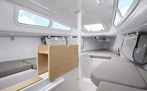 Intérieur du FIRST 24, cabine lumineuse avec banquettes grises, table en bois et hublots ouverts sur le ciel bleu.