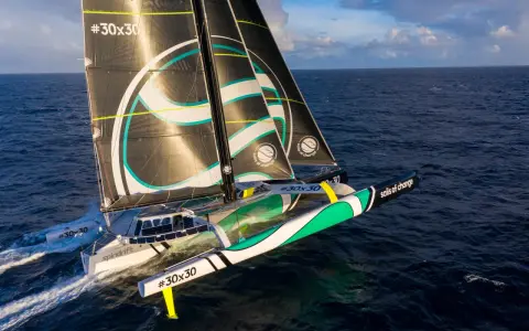Un trimaran aux voiles noires et blanches, arborant le logo "sails of change", fend les vagues bleues sous un ciel nuageux.