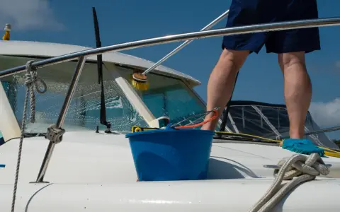 Un homme effectue l'entretien de bateau, lavant la proue avec une brosse et un seau bleu.