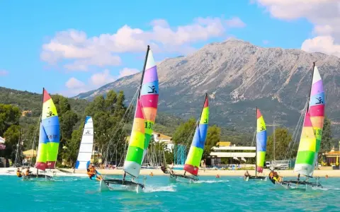 Plusieurs Hobie Cat 16 naviguent sur une mer turquoise, voiles multicolores, montagne en arrière-plan.