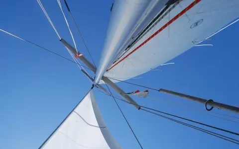 Vue plongeante sur le mat de bateau, voiles blanches déployées sous un ciel bleu éclatant, cordages tendus.