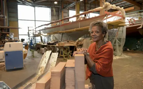 Jacqueline Tabarly, souriante, dans un atelier de construction navale, devant des blocs de bois et un voilier en cours de fabrication.