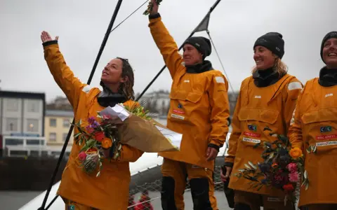 Des femmes en cirés jaunes célèbrent la fin du célèbre projet, bouquets à la main.