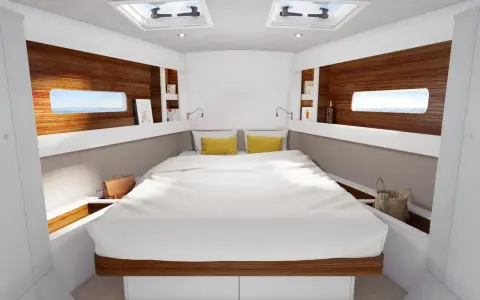 L'intérieur d'un voilier 8m, avec un grand lit blanc, des coussins jaunes, des hublots donnant sur la mer et des étagères en bois.