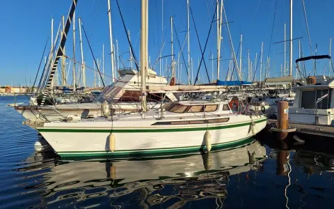 Un voilier blanc et vert, un Jeanneau Espace 1000, reflète sur l'eau calme d'un port.
