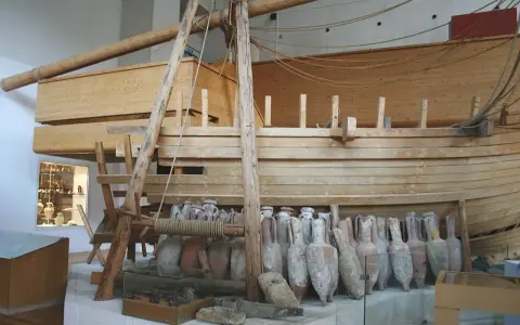 Amphores antiques alignées devant une reconstitution de navire en bois, rappelant le transport de vin par un ancien pinardier.