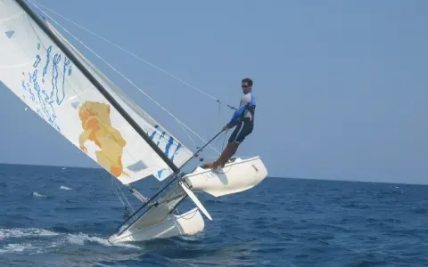Un homme en trapeze sur un catamaran navigue sur une mer bleue sous un ciel clair. La voile arbore un dessin original.