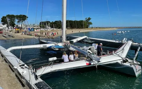 Le trimaran Flo, un bateau à trois coques, est amarré à quai. Des personnes sont à bord et sur la plage en arrière-plan.
