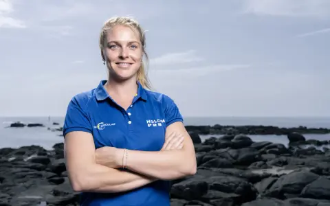 Rosalin Kuiper, souriante, pose les bras croisés devant un paysage côtier rocheux.