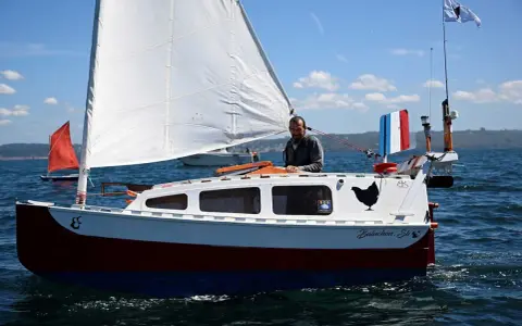 Un homme sourit sur son bateau nommé "Baluchon". Voiles blanches, coque rouge et bleue, drapeau français.