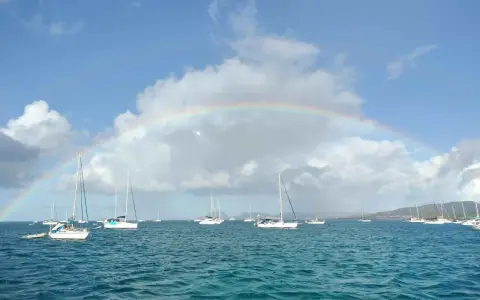 Un arc-en-ciel sur une mer bleue, parsemée de voiliers. L'appel de l'aventure : tout quitter pour vivre sur un bateau.
