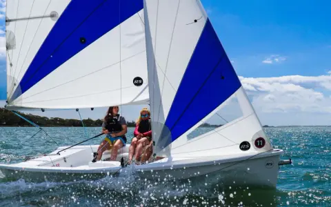 Deux jeunes femmes naviguent sur un voilier RS Venture par une journée ensoleillée. L'eau scintille sous le ciel bleu.