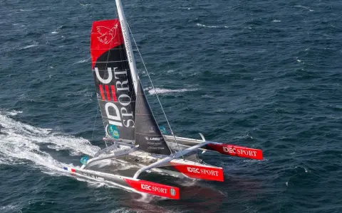 Le trimaran IDEC SPORT fend les vagues, une voile rouge et noire ornée d'un oiseau blanc.