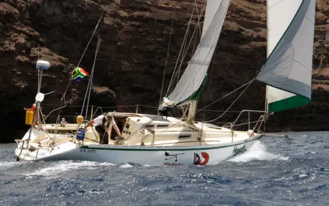 Un voilier cap horn, le "Windicator", navigue près d'une côte rocheuse. Le drapeau sud-africain flotte fièrement.