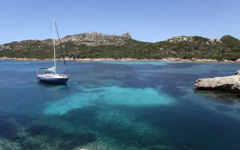 Un voilier bleu ancré dans les eaux turquoise de Cap Corse, avec des collines verdoyantes en arrière-plan sous un ciel clair.