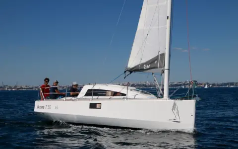 Un voilier Ikone 7.50 navigue par beau temps, avec trois personnes à bord. La voile est hissée, le ciel est bleu.