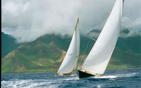Deux voiliers de course, dont un avec le numéro 7020, naviguent sur une mer agitée, avec des montagnes verdoyantes et brumeuses en arrière-plan. Le pen duick 3 est en pleine action.