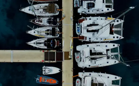 Vue aérienne d'un port avec plusieurs yachts à voile blancs amarrés, certains catamarans, et des annexes gonflables.