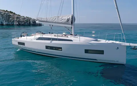 Un voilier blanc moderne navigue sur des eaux turquoise claires, près d'une île rocheuse. Ce modèle, le "Oceanis 42", pourrait bien être le meilleur voilier habitable pour vos aventures.