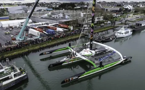 Le trimaran Sodebo Ultim 3, prêt pour la course, est amarré dans un port, entouré de spectateurs et de grues.